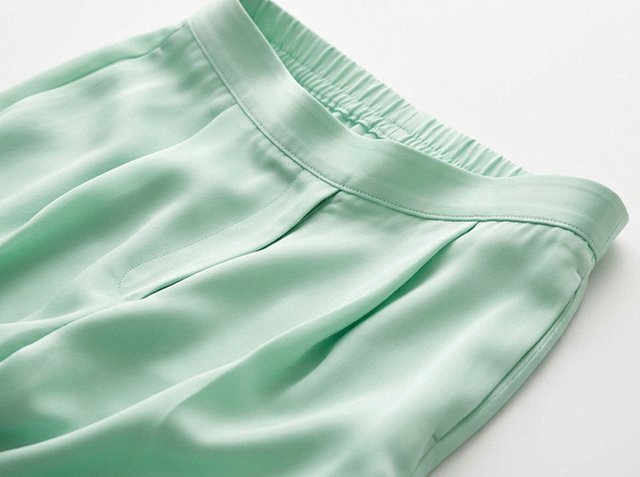 Pantaloni in raso verde chiaro con vita elastica e pieghe frontali stesi su una superficie bianca.
