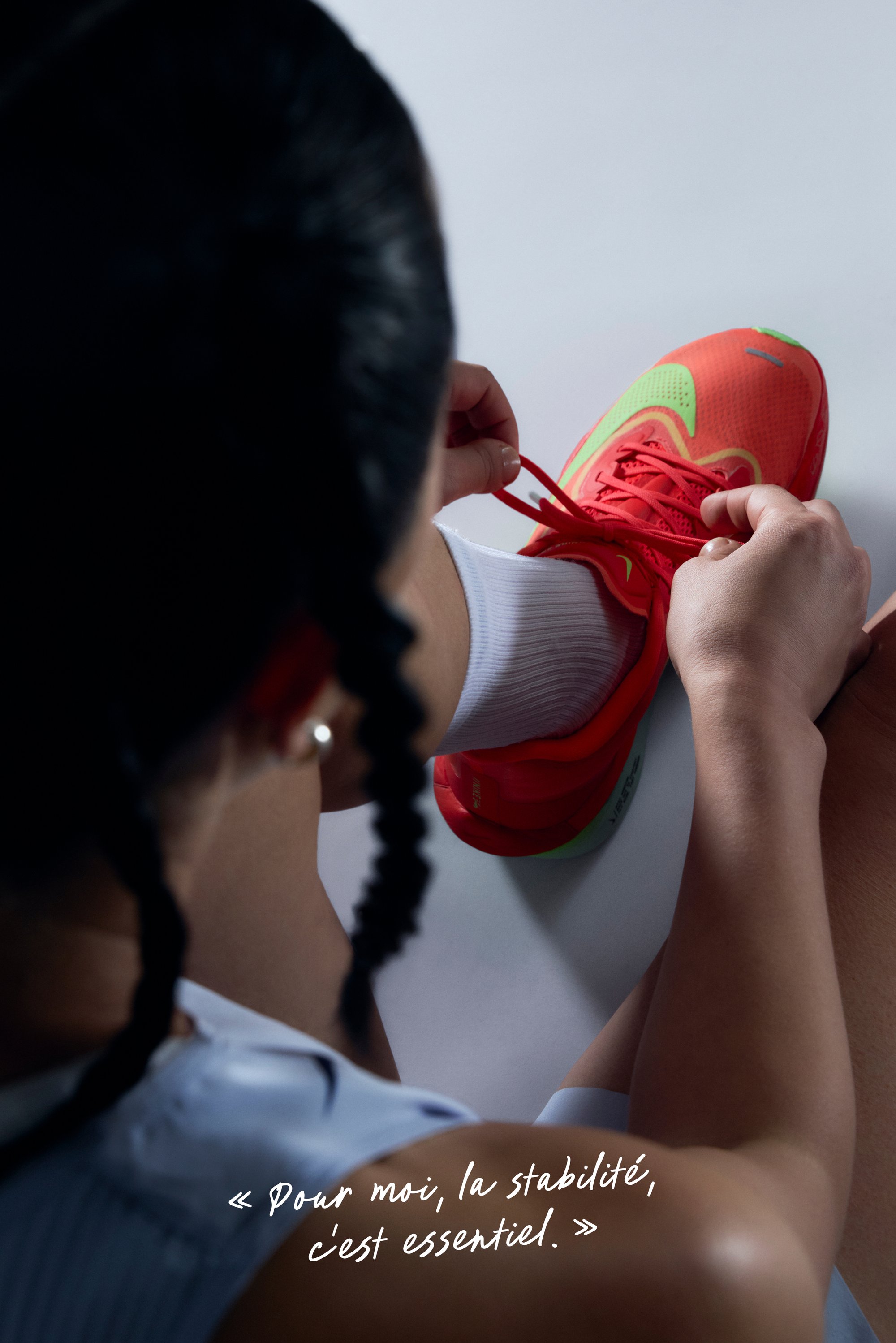 Bottine de sport rouge vif avec des accents verts et une tige en maille texturée, en train d'être nouée par une personne portant des chaussettes blanches, sur un fond neutre.