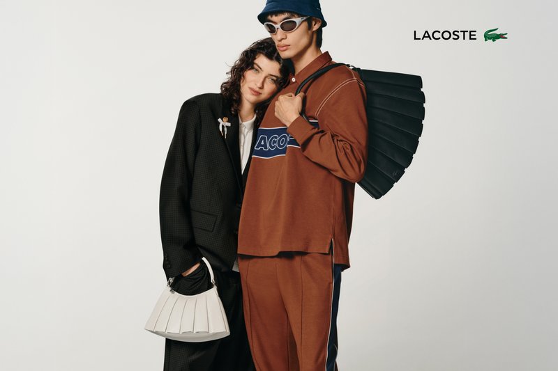 Deux mannequins posent de manière rapprochée, l'un portant un costume noir et l'autre un haut orange arborant la marque "LACOSTE", tenant des sacs élégants sur fond uni.