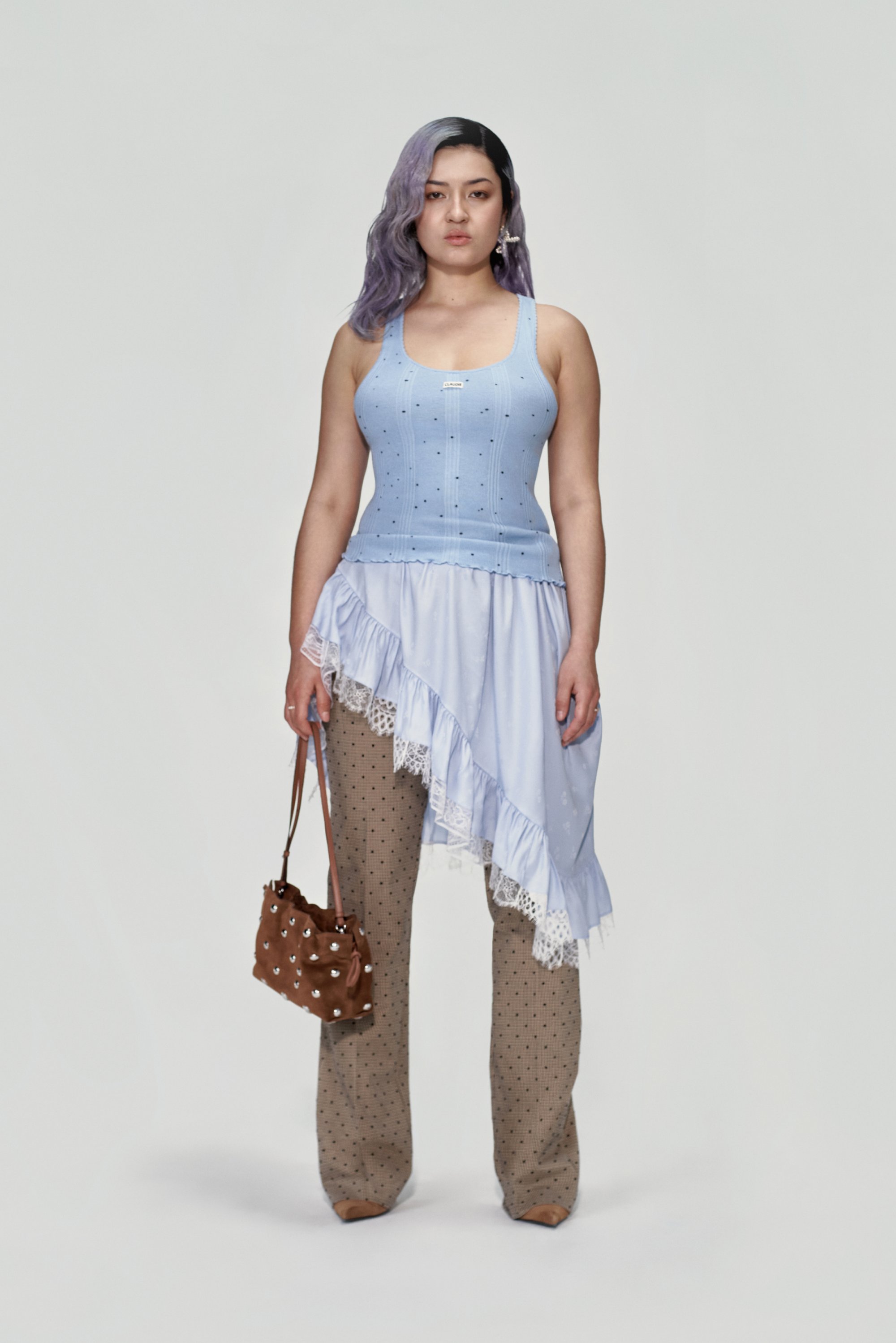 Una giovane donna con i capelli viola indossa una canotta blu e un vestito a strati, mentre tiene in mano una borsa marrone, su uno sfondo neutro.