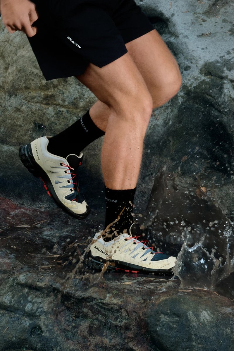 Scarpe outdoor beige e nere con lacci rossi e suola testurizzata, spruzzando acqua su una superficie rocciosa. Design leggero per agilità.
