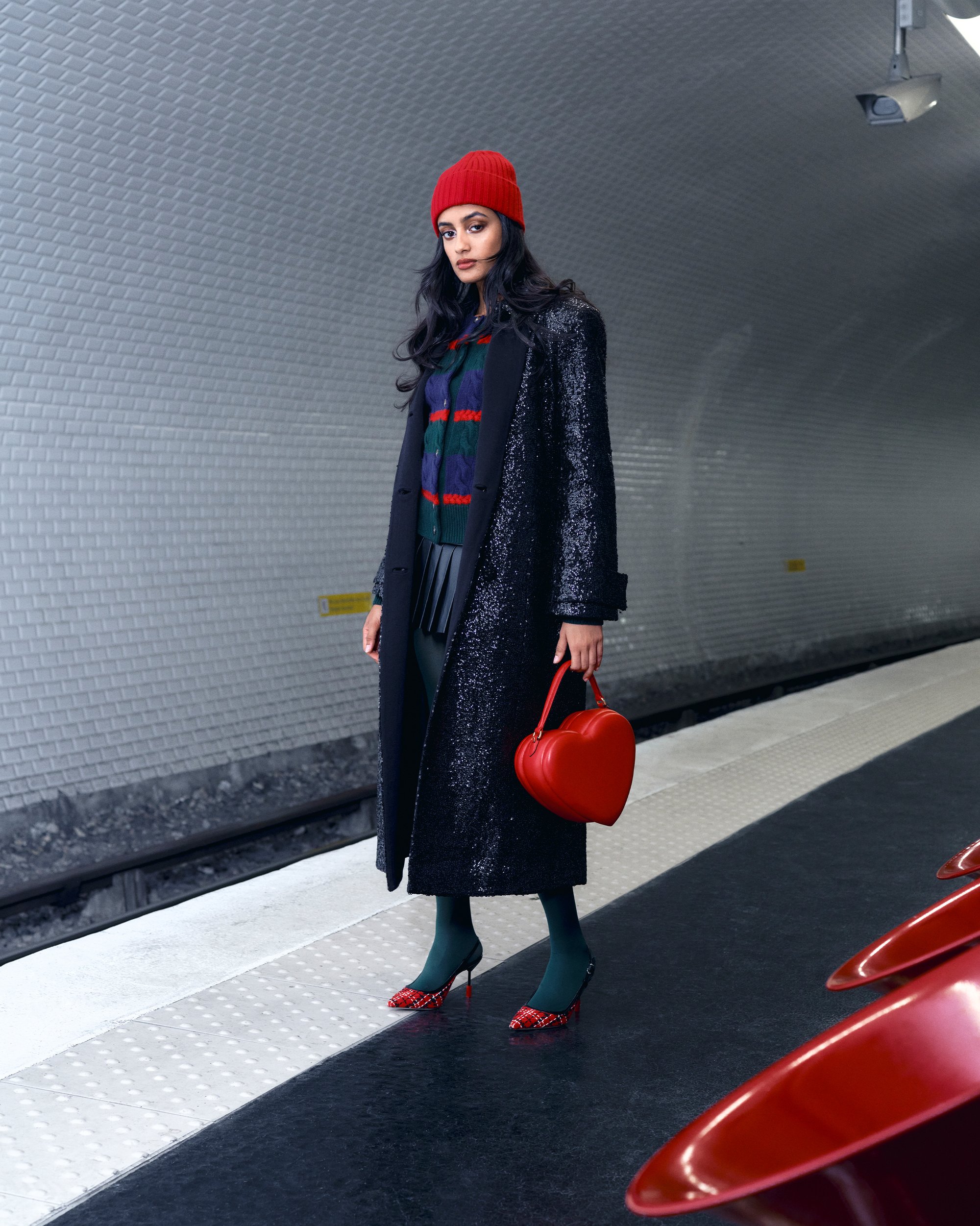 Een vrouw staat op een metroplatform, gekleed in een rode beanie, een sprankelend zwart jasje en een hartvormige rode handtas, met gemusterde hakken.