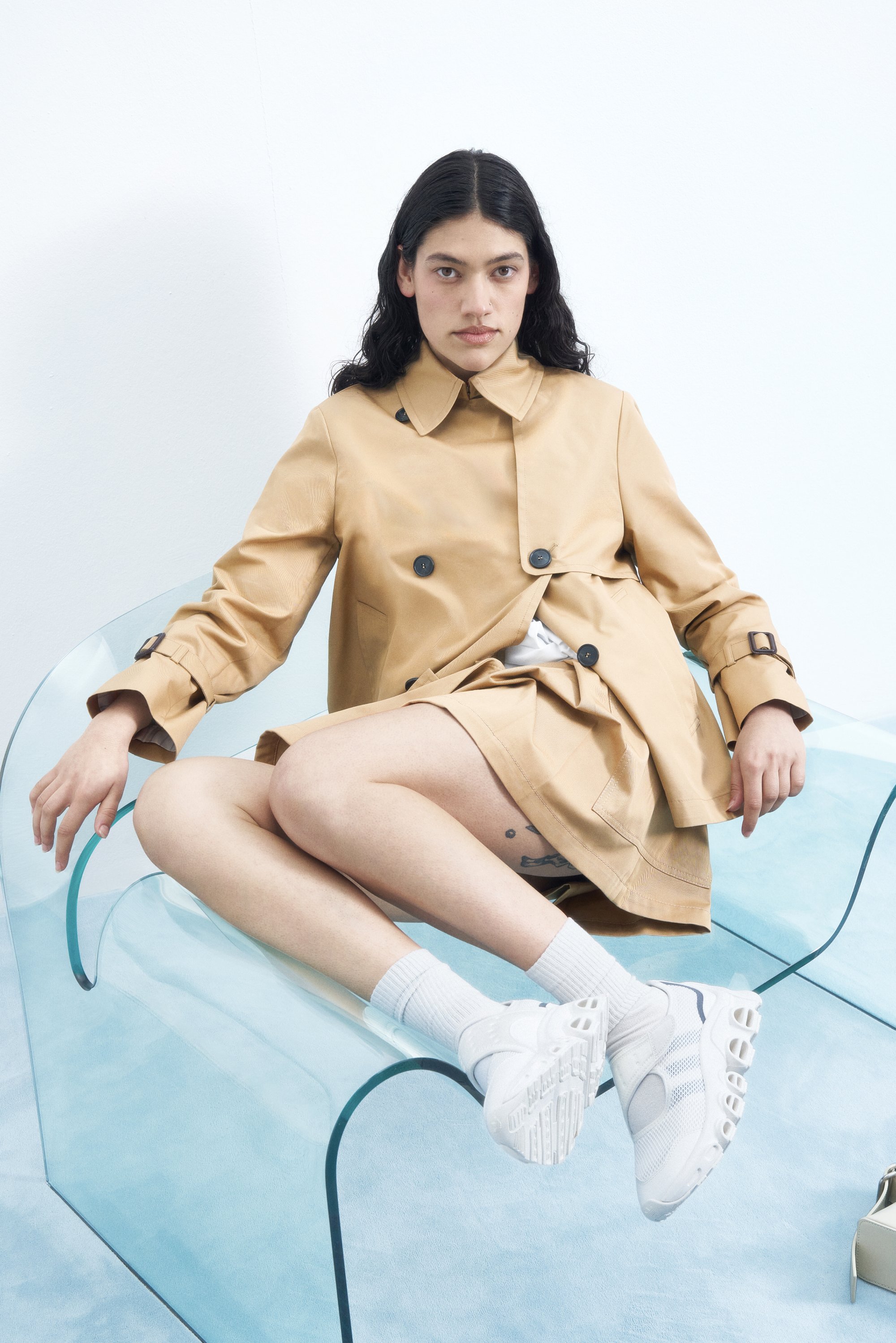 Una persona con el cabello largo y oscuro está sentada en una silla transparente, vestida con un abrigo trench beige, calcetines blancos y zapatillas ligeras, con un fondo minimalista.