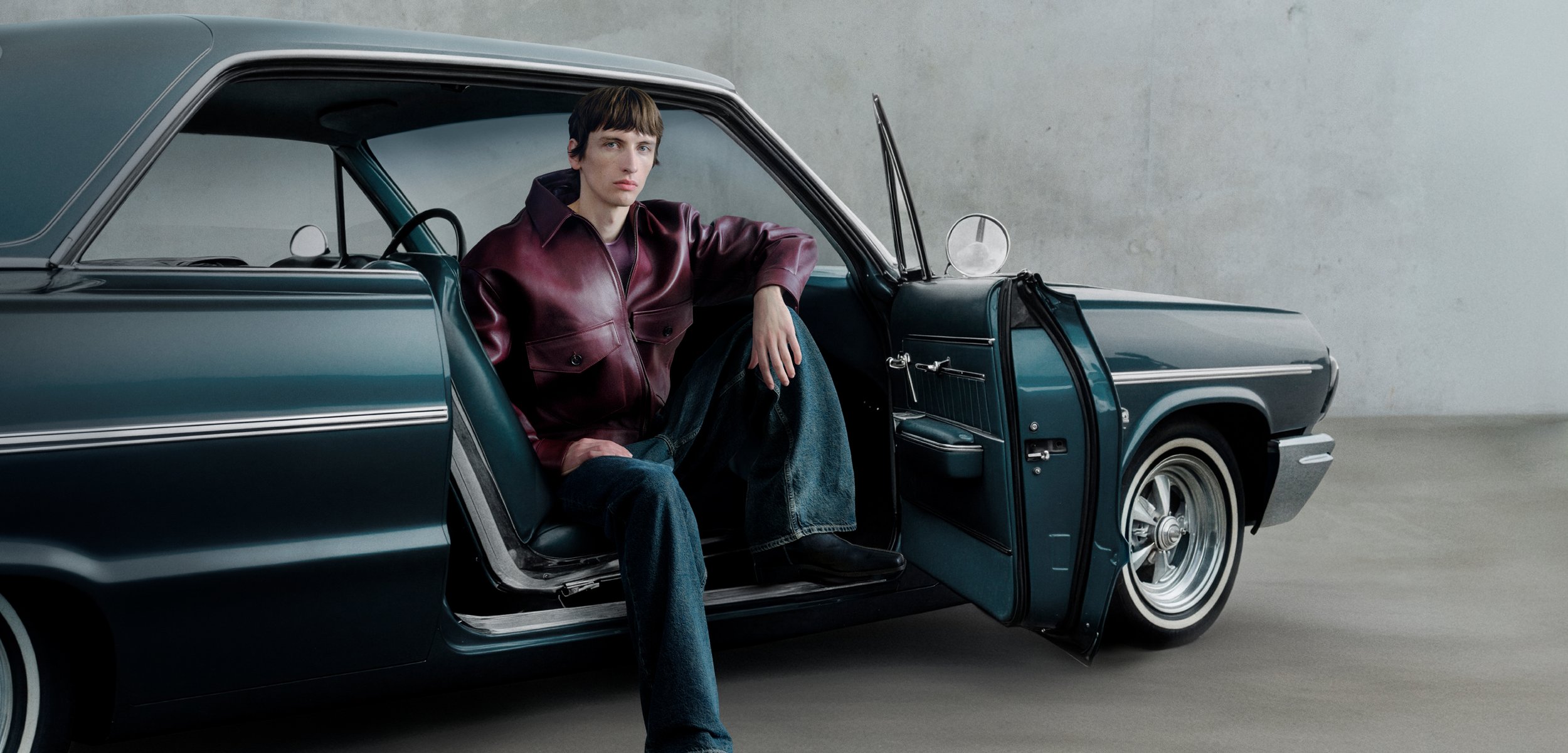 Vintage blue car with chrome accents, door open; person wearing a maroon leather jacket and denim seated inside in a relaxed posture.