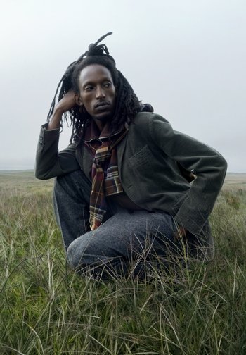Modèle assis dans l'herbe, portant un blazer vert en velours côtelé, un pantalon gris texturé et une écharpe à carreaux, avec de longues dreadlocks et une expression pensive.