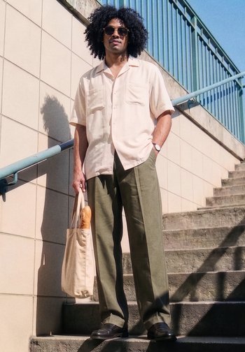 Homme aux cheveux bouclés et lunettes de soleil tient un sac fourre-tout avec une baguette, debout sur des marches extérieures à côté d'un mur carrelé et d'une rambarde en métal.