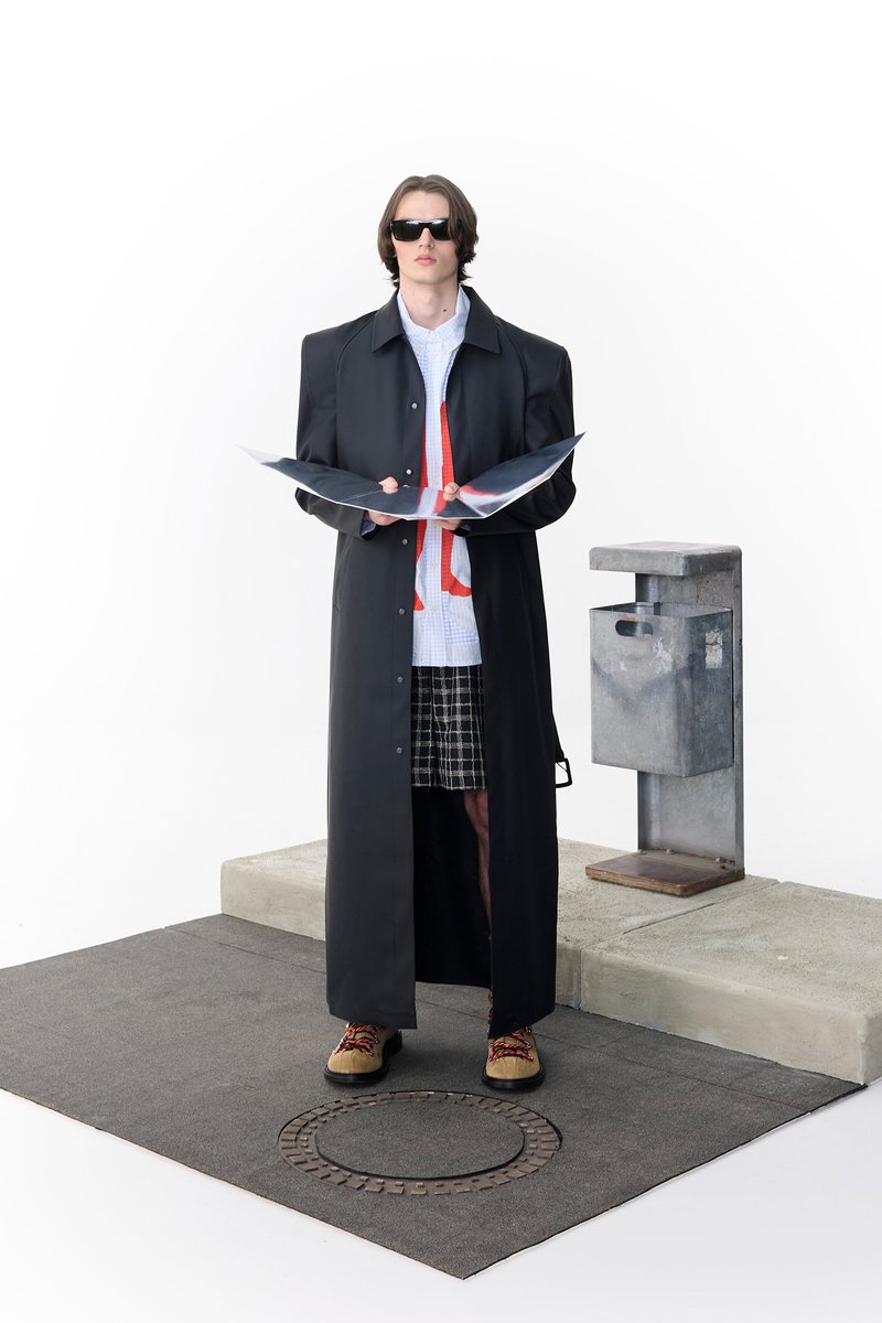 A person in oversized black outerwear and sunglasses holds open fashion magazines next to a grey mail receptacle on a textured surface.