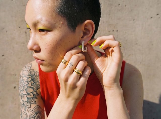 Personne aux cheveux courts et tatouage ajuste une boucle d'oreille géométrique, portant un haut sans manches rouge, du vernis à ongles jaune et des bagues en or.