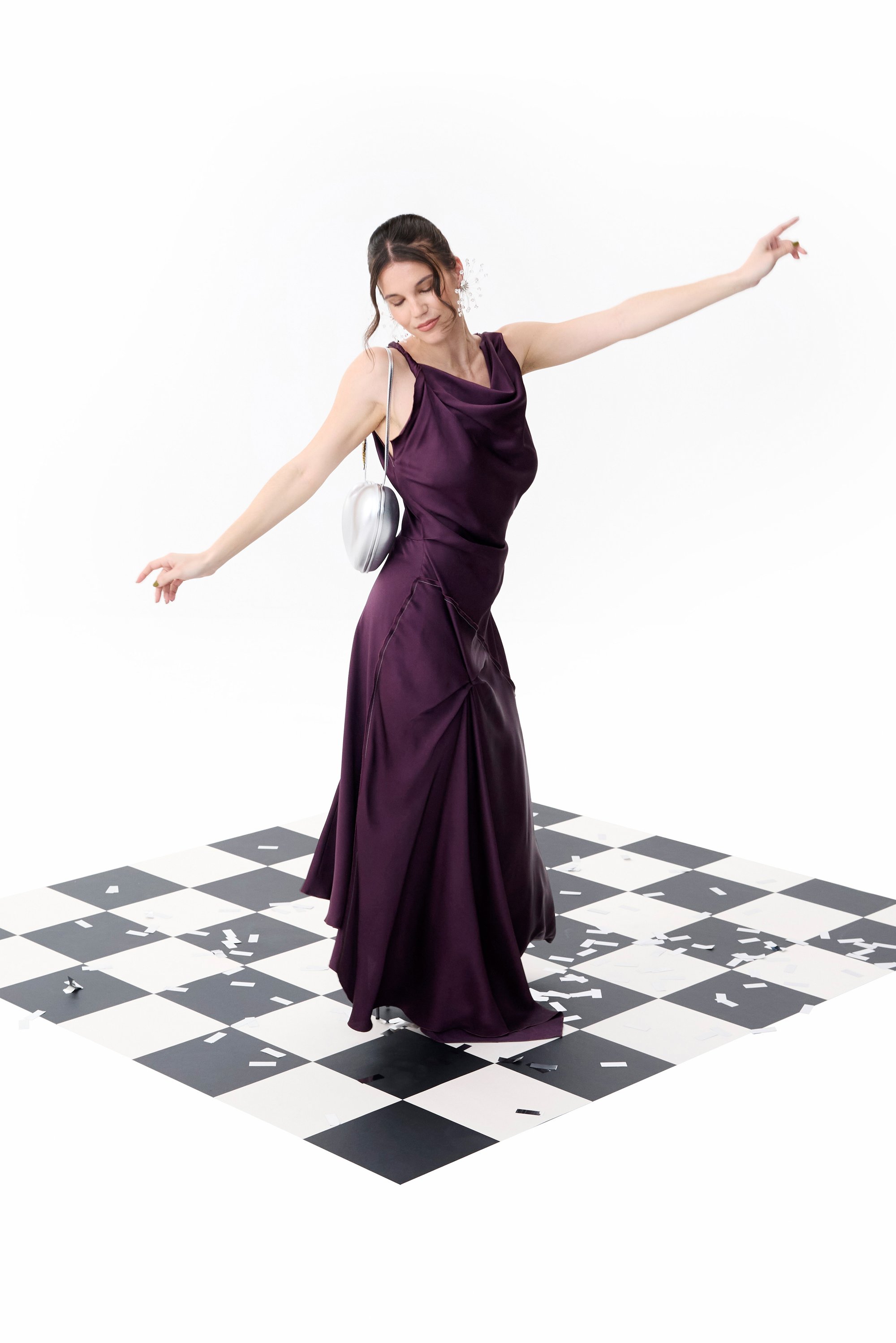Una mujer con un vestido púrpura fluido baila sobre un suelo de cuadros, rodeada de confeti, irradiando alegría y elegancia.