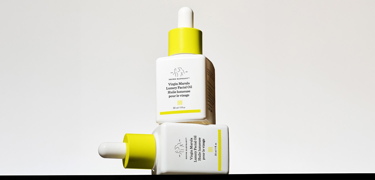 Two white dropper bottles with yellow caps labeled Virgin Marula Luxury Facial Oil, one standing upright and one lying horizontally.