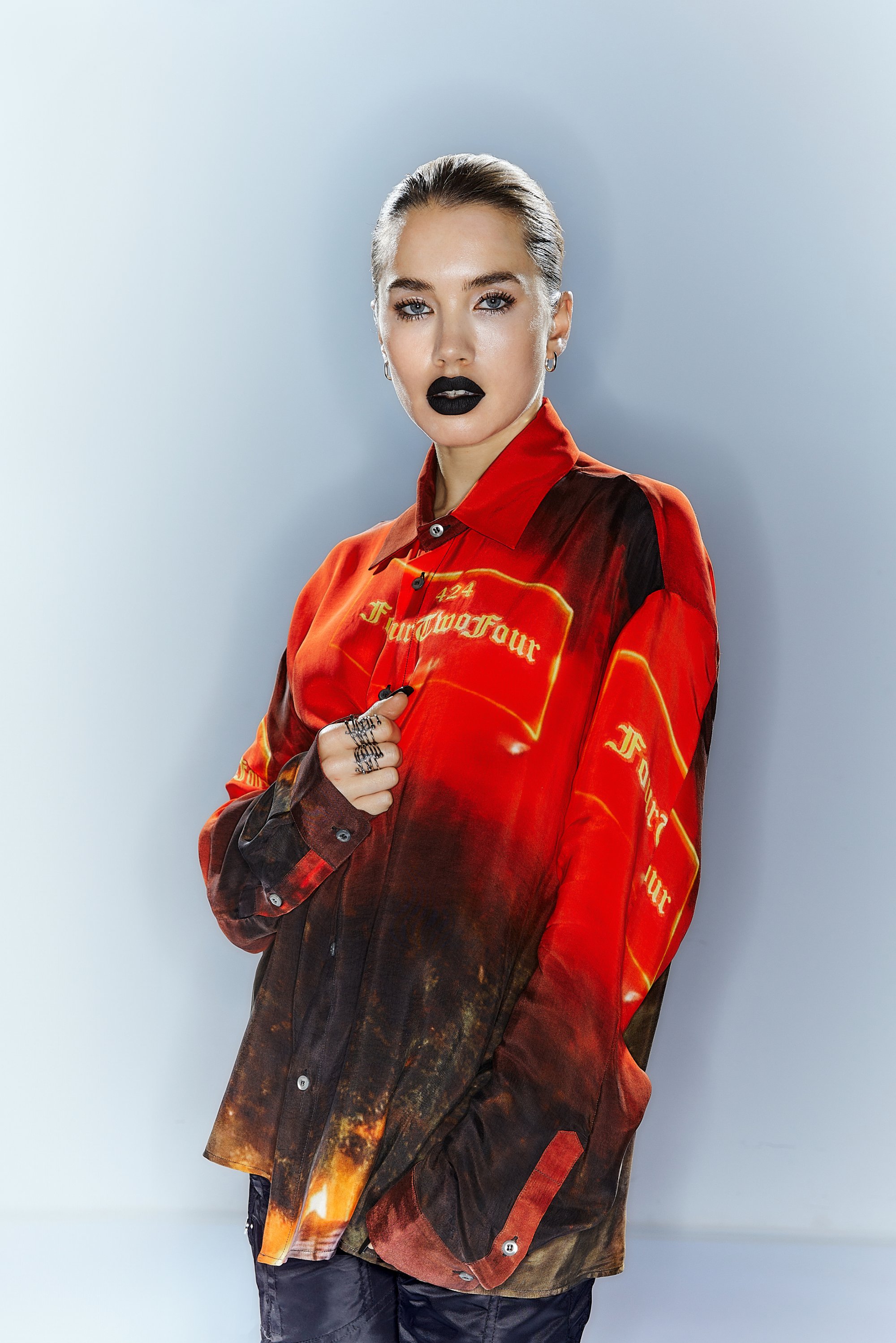 Een vrouw met naar achteren gekamd haar en zwarte lippenstift poseert in een levendig rood en zwart shirt, terwijl ze zilveren ringen om haar vingers toont.
