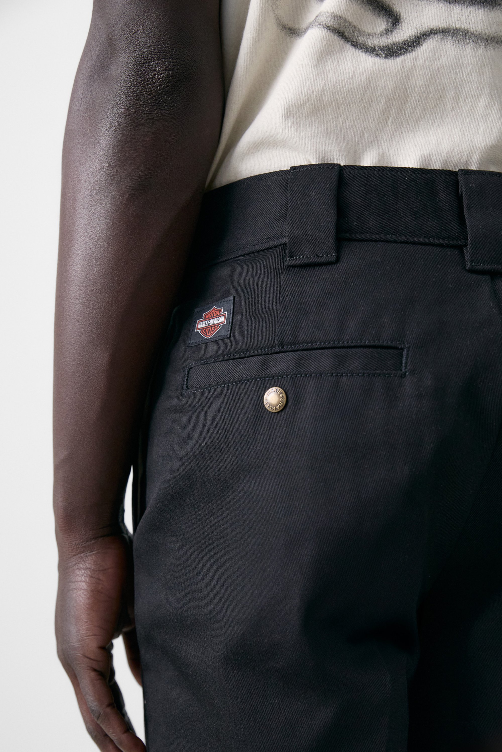 Een close-up van de arm en heup van een persoon, die zwarte broek met een Harley-Davidson logo en een gouden knoopdetail toont.