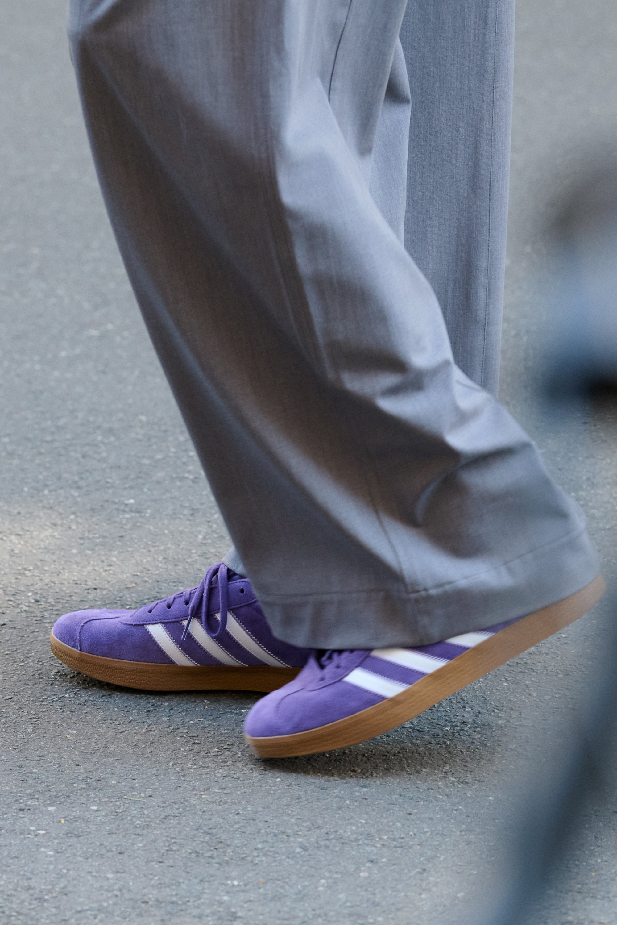 Un pie que lleva zapatillas moradas con rayas blancas está pisando un pavimento gris, acompañado de pantalones grises holgados.