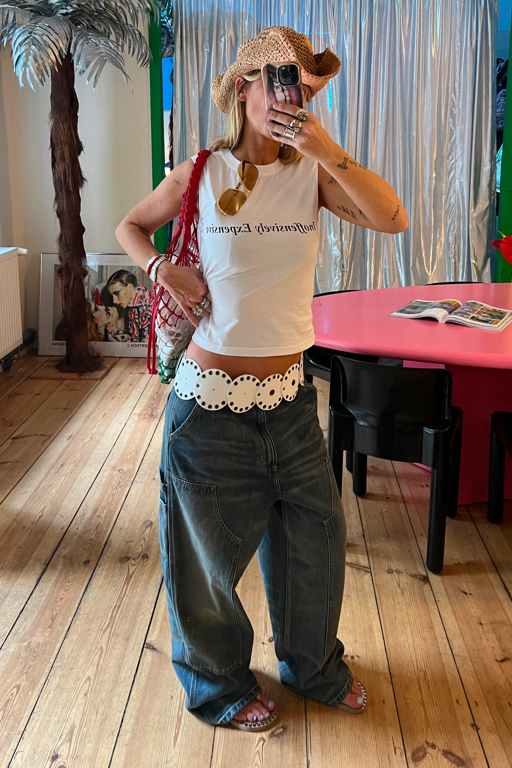 Mujer con jeans anchos, cropped blanco con texto, cinturón blanco grande, sombrero de paja y bolso rojo tomando un selfie frente al espejo en una habitación colorida con mesa rosa.
