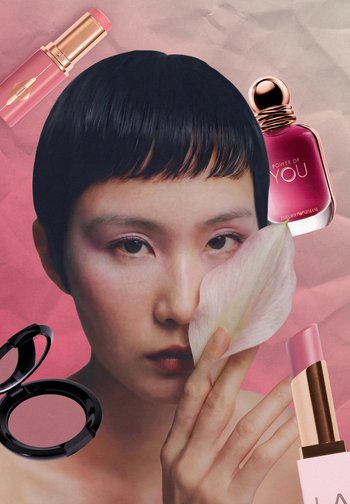 Donna con capelli corti e neri tiene un petalo di fiore bianco vicino al viso, circondata da prodotti makeup rosa e una boccetta di profumo.