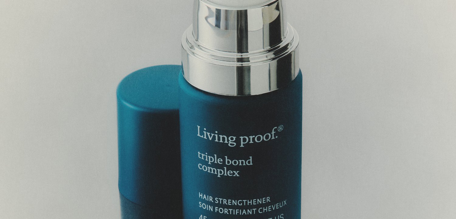 Niebieska cylindryczna butelka z srebrną pompką oznaczona Living proof triple bond complex hair strengthener. Kapturek umieszczony za butelką na białej powierzchni.
