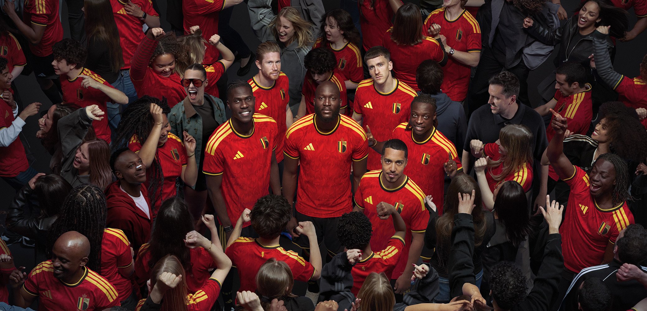 Groep diverse voetballers in rode en gele Belgische shirts die zelfverzekerd staan temidden van juichende fans die de teamkleuren dragen.