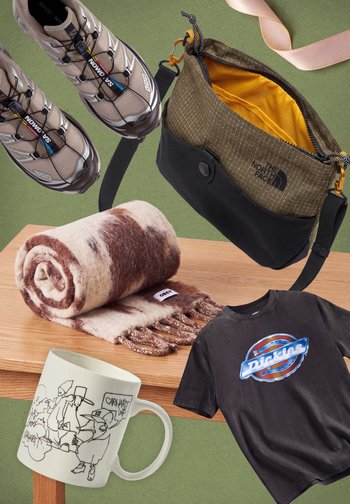 Des articles de plein air et de streetwear disposés sur une table : des chaussures de randonnée, un sac North Face ouvert, une écharpe à motifs, un T-shirt à imprimé graphique, un mug illustré et un ruban rose.
