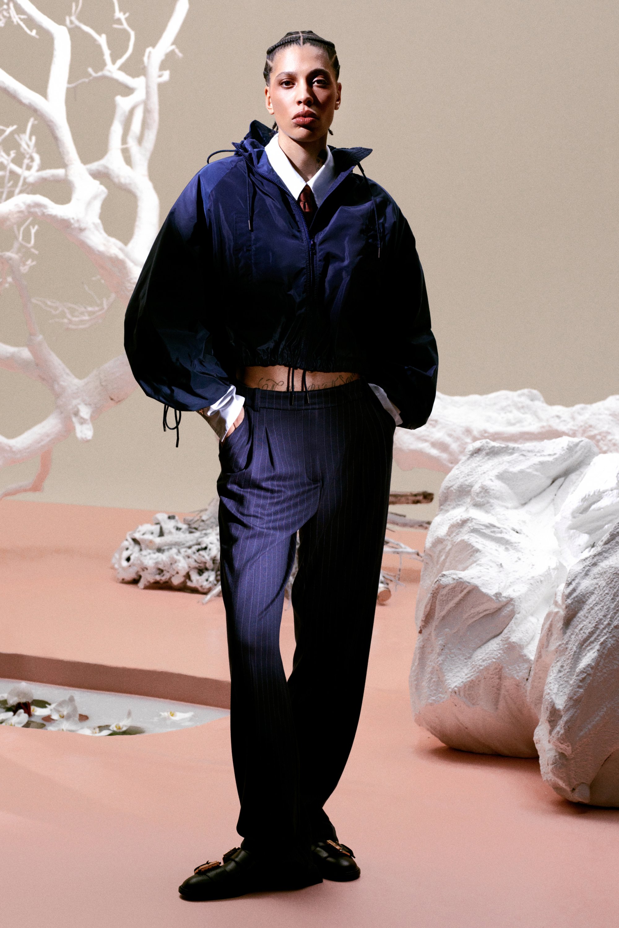 Una modelo de moda se presenta con confianza en una chaqueta cortada marina y pantalones de rayas, con un fondo minimalista de ramas blancas y rocas.