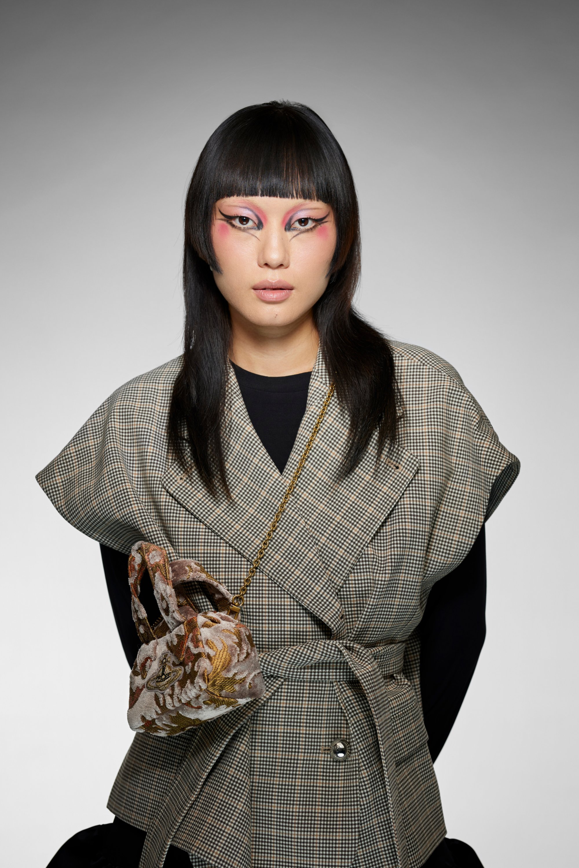 A model with long, dark hair wears a plaid wrap top and showcases a small patterned handbag. Bold, artistic eye makeup enhances her look.