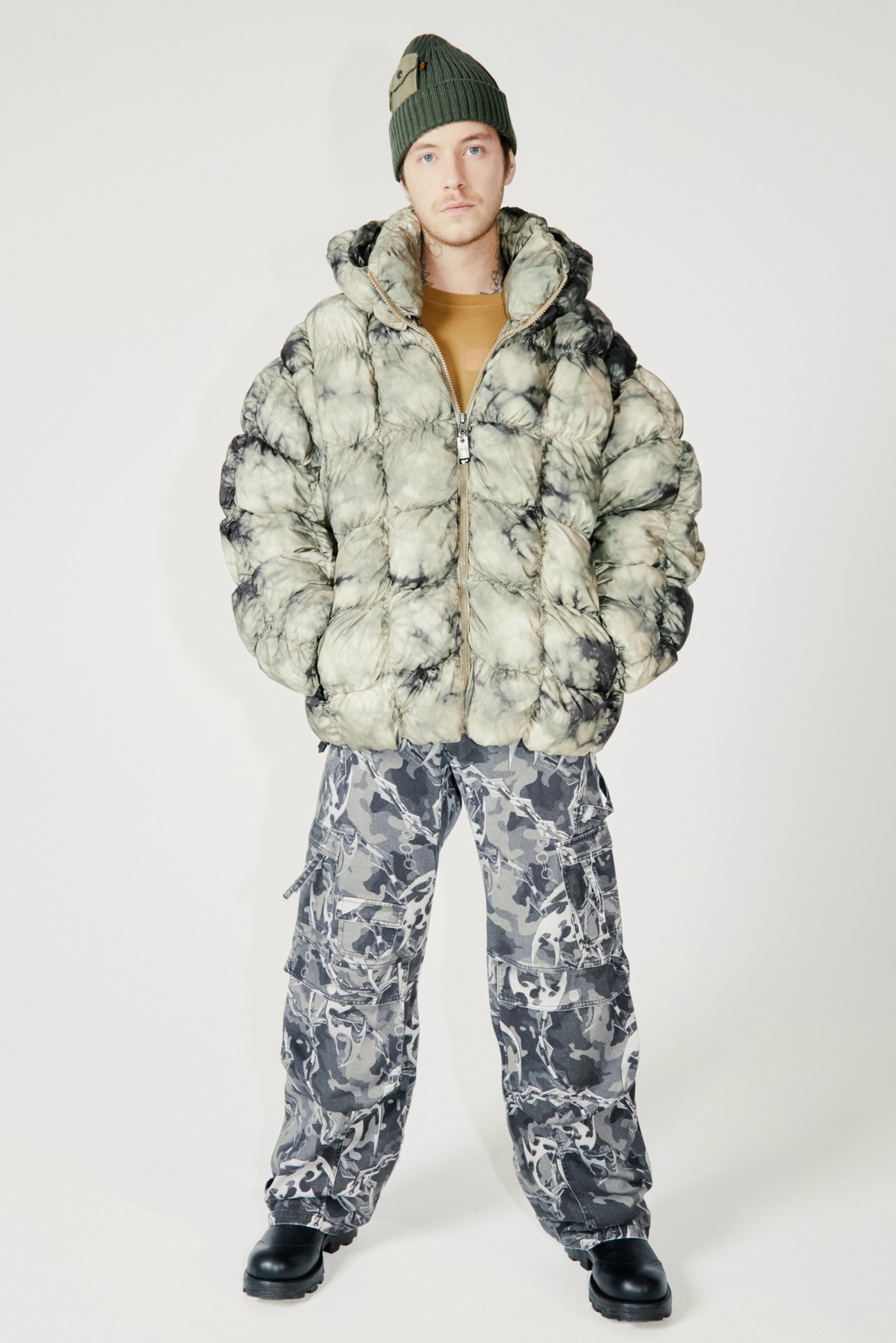 Una persona está de pie con una chaqueta de tie-dye esponjosa y pantalones de cargo, usando un sombrero beanie verde. El fondo es completamente blanco, lo que resalta el atuendo.