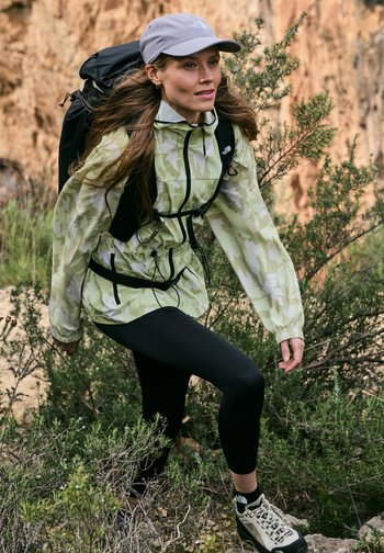 Woman hiking uphill wearing a light green camouflage jacket, black leggings, white hat, and backpack in a rocky, shrub-filled outdoor area.
