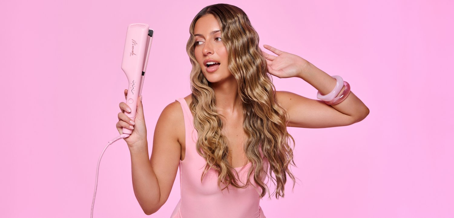 Femme aux longs cheveux ondulés tient un fer à lisser rose et fait un geste de l'autre main devant un fond rose.