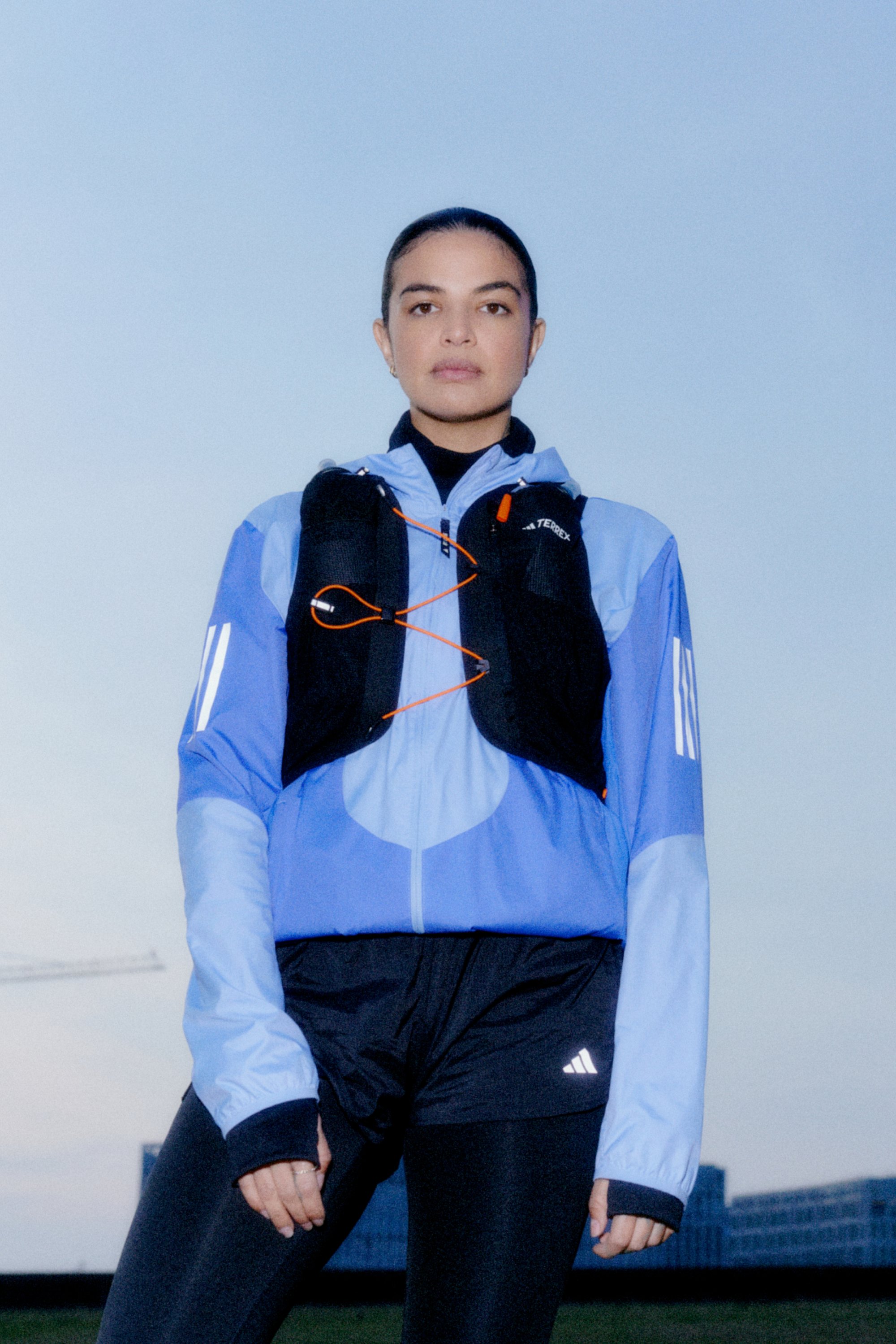 Mujer atlética vestida con una chaqueta azul y un chaleco negro posa con confianza contra un cielo despejado, con edificios urbanos al fondo.