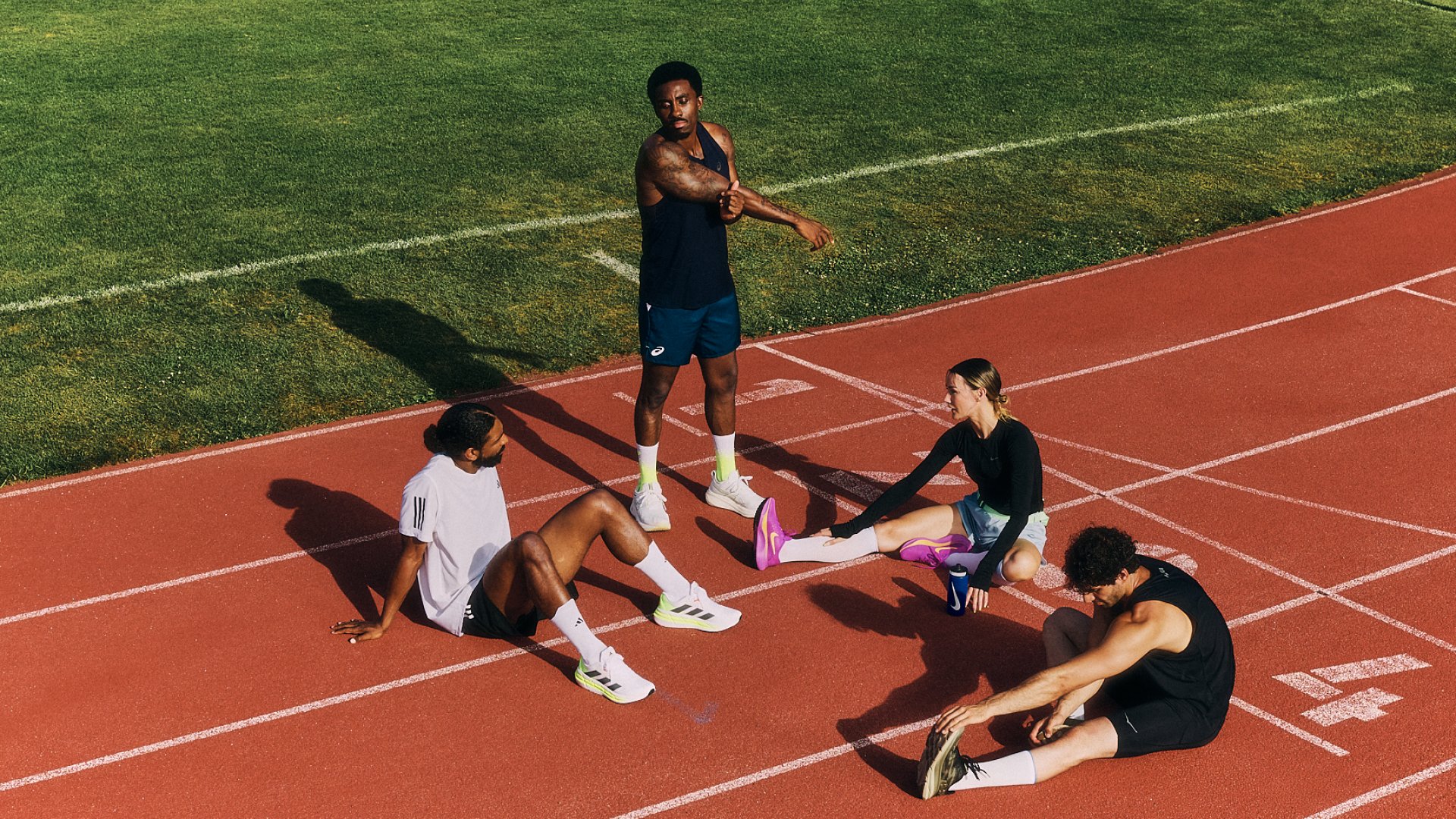 Four athletes stretch on a track field. One stands, demonstrating an exercise, while three sit, focused on warming up. Green grass surrounds them.
