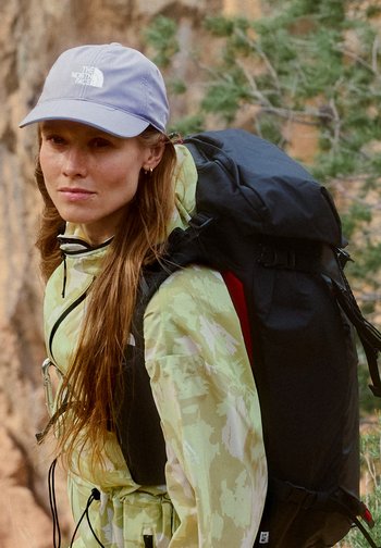 Vrouw met lang haar, gekleed in een licht camouflagejack en een lavendelpet, draagt een grote zwarte rugzak voor wandelen in een bosrijke omgeving.