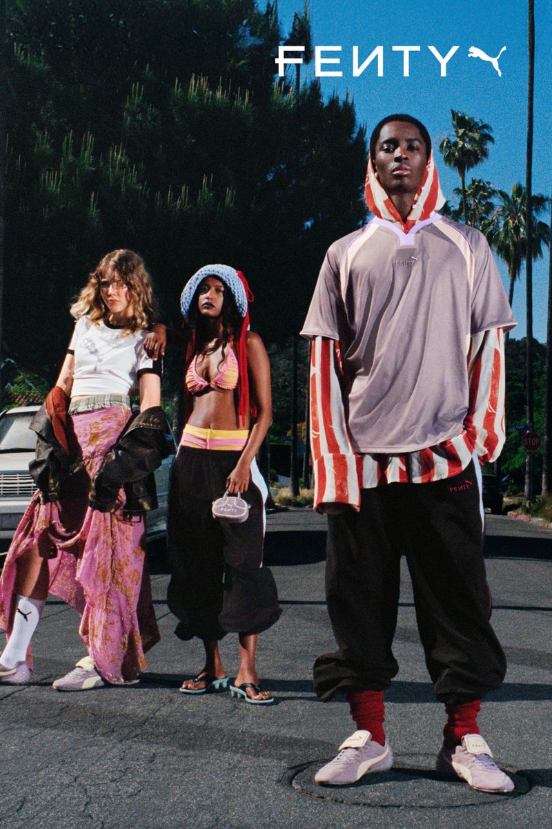 Tres modelos están de pie en una calle vistiendo ropa de FENTY X PUMA. Las características destacadas incluyen una falda estampada rosa, tops con capucha y pantalones deportivos coloridos.
