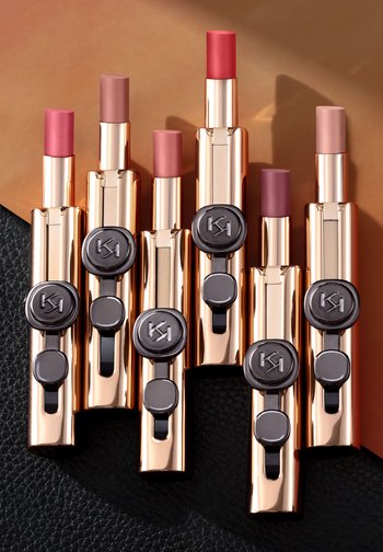 Six lipsticks in gold metallic tubes with black accents. Shades range from deep pink to nude. Smooth finish, varying heights, and logo detail.
