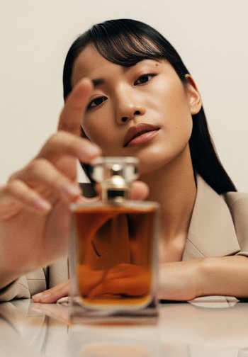 Perfume bottle with a square glass base and a silver cap. The liquid is amber-colored. A hand is reaching toward the bottle.
