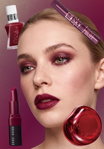 Gros plan sur un mannequin portant un maquillage des yeux et un rouge à lèvres bordeaux, entouré de vernis à ongles rouge, de mascara, de rouge à lèvres et d’un boîtier cosmétique rond et rouge.