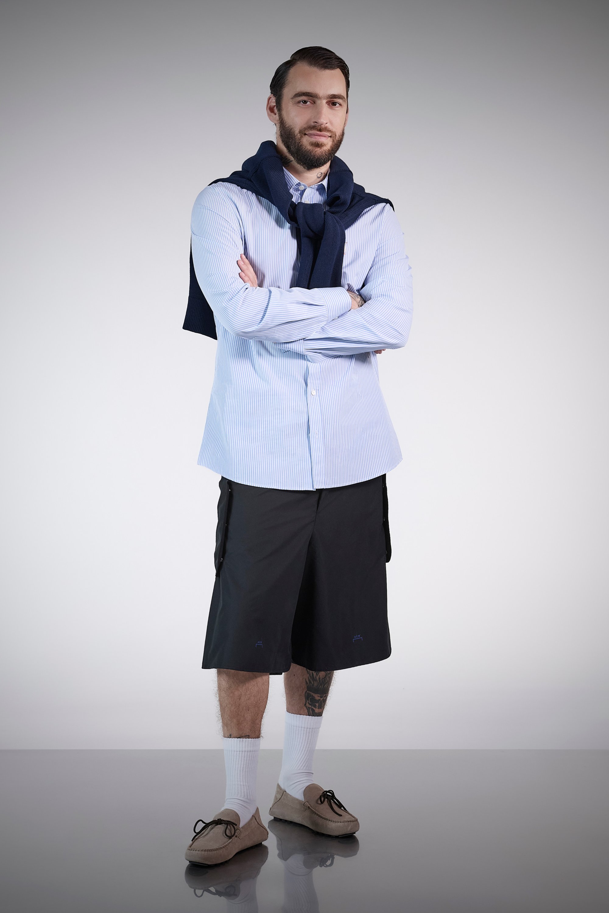 A man stands confidently with his arms crossed, dressed in a blue striped shirt, black shorts, and light shoes, with a sweater draped around his shoulders.