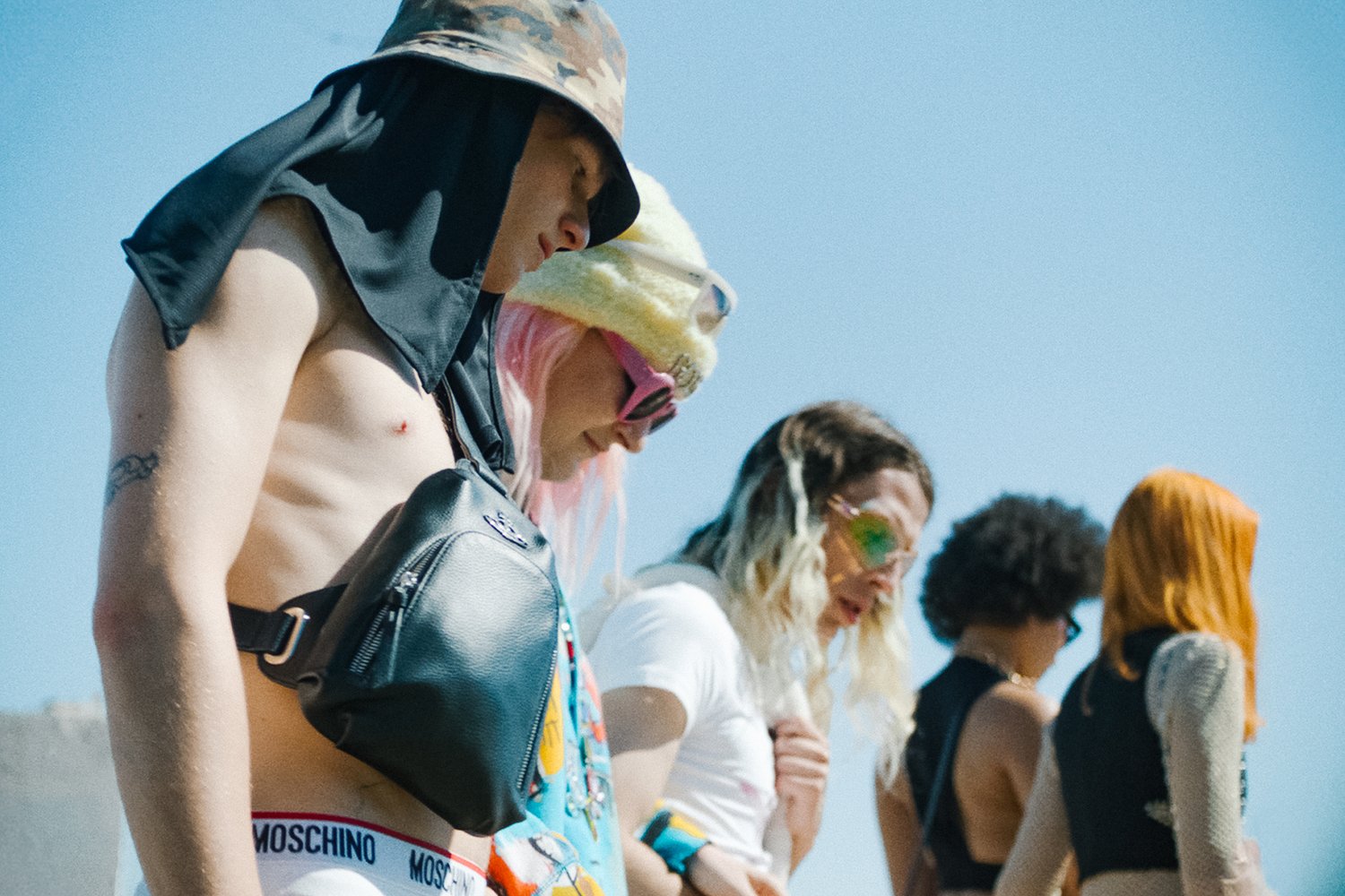 Vijf individuen in verschillende stijlen, waaronder levendige haarkleuren en accessoires, lopen samen onder een heldere blauwe lucht en richten hun aandacht geconcentreerd vooruit.