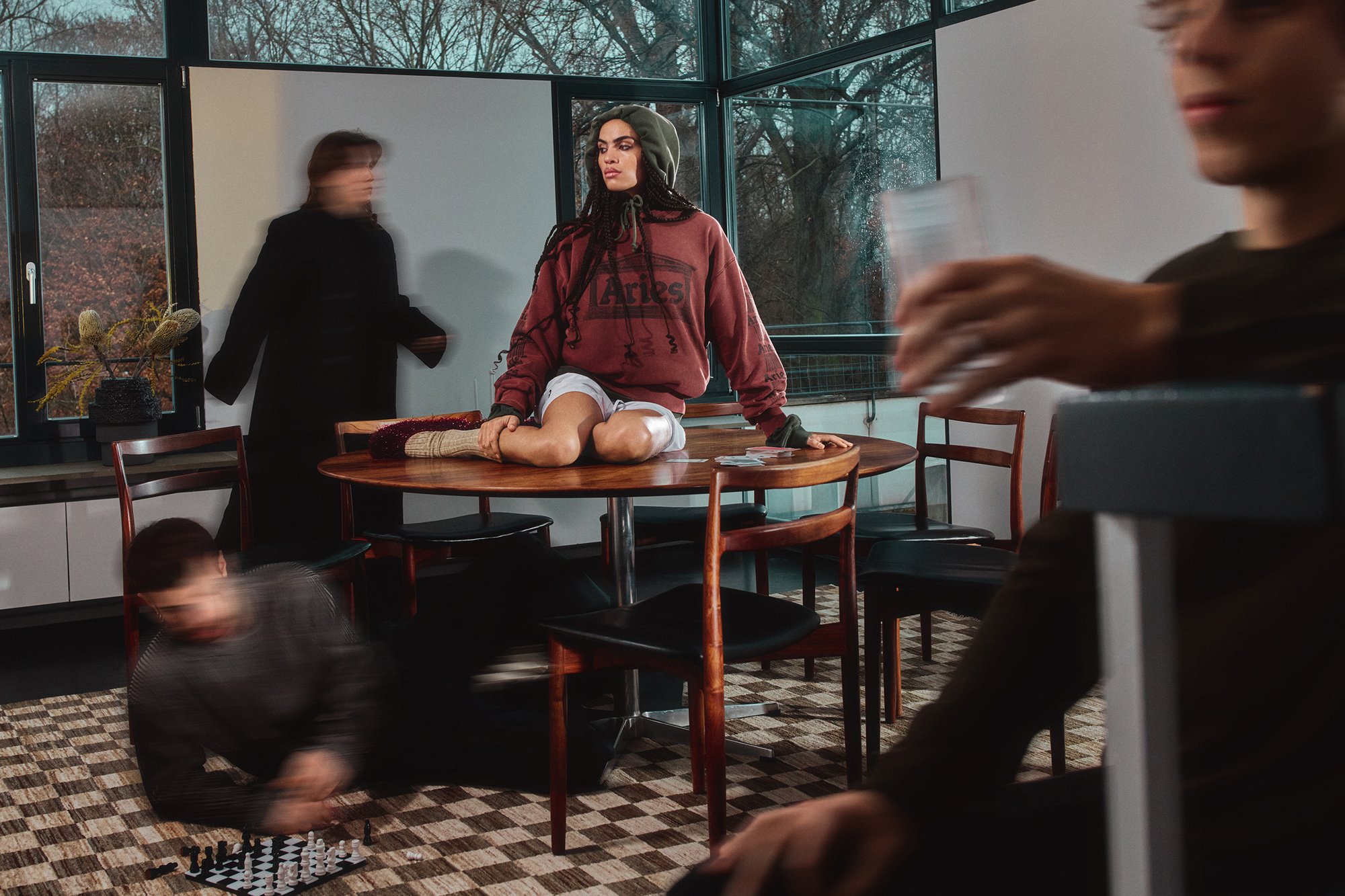 Una persona con capelli lunghi è seduta a un tavolo, indossando un felpa e pantaloni corti. Due figure sfocate si muovono nella stanza, con pezzi di scacchi sul pavimento.