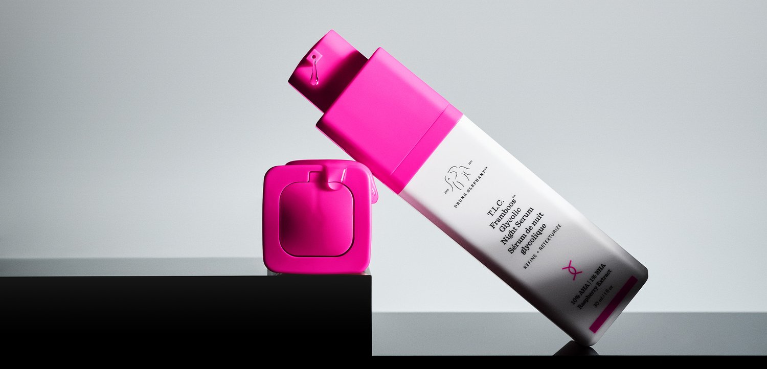 Eine weiß-pinke Drunk Elephant T.L.C. Framboos Glycolic Night Serum Flasche lehnt an ihrem passenden Deckel auf einer glänzenden schwarzen Oberfläche.