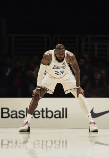 Un joueur de basketball en tenue blanche des Lakers est accroupi intensément sur le parquet, son regard dirigé vers l'avant, avec un public visible en arrière-plan.
