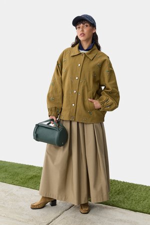 Olivgrüne Jacke mit Blumenstickerei, brauter Rock, braune Blockabsatzstiefel und eine grüne zylindrische Tasche. Baseballkappe auf dem Kopf.