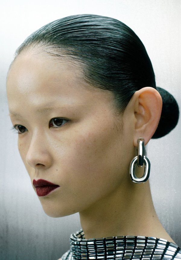 Close-up of a person with slicked-back hair, wearing large silver chain-style earrings and a structured metallic collar.