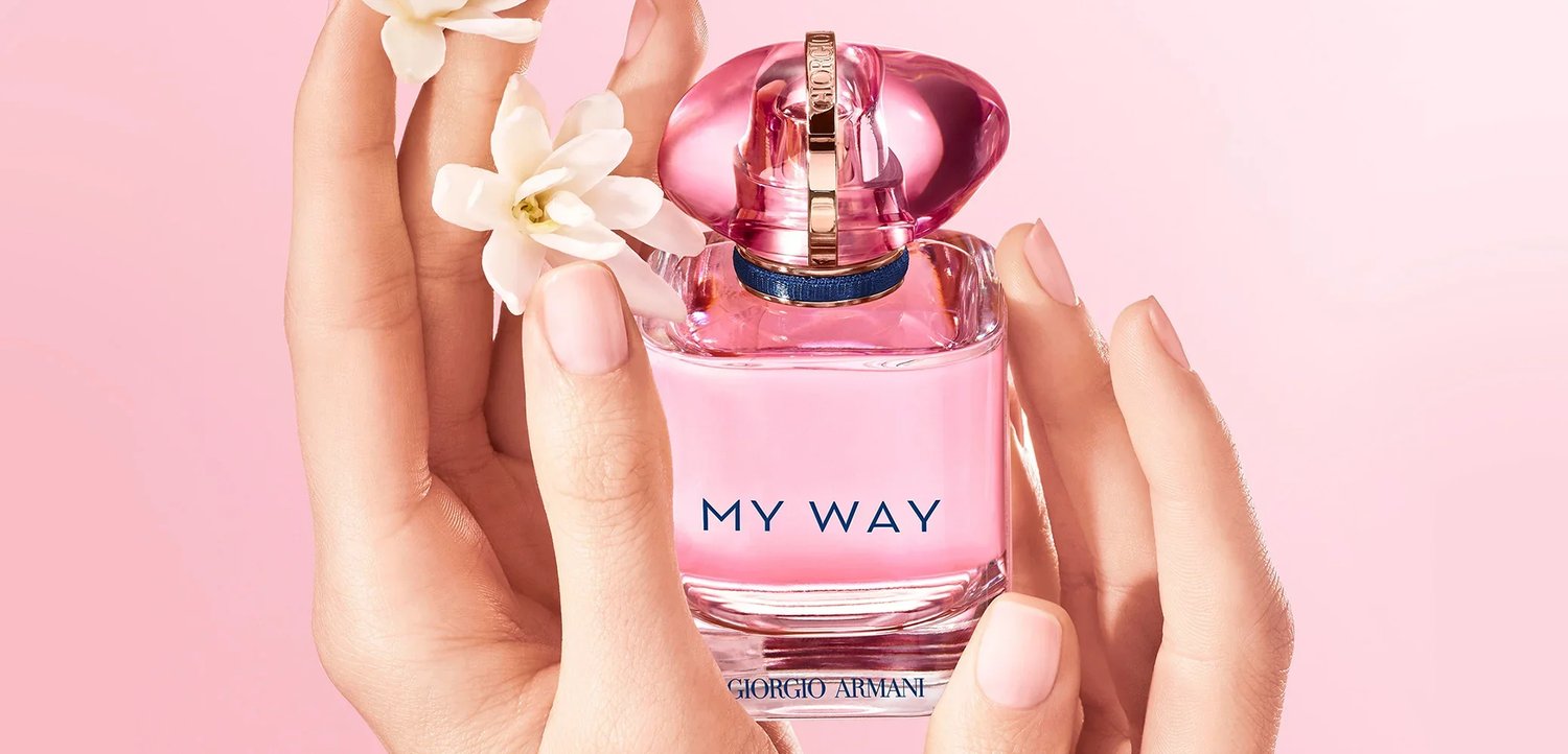Mains aux ongles rose pâle tenant un flacon de parfum rose "My Way" de Giorgio Armani orné de petites fleurs blanches sur un fond rose doux.