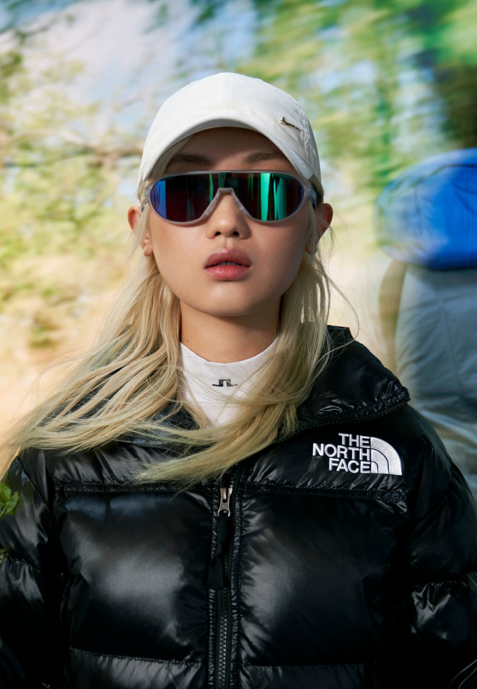 Eine Person mit langen blonden Haaren trägt eine reflektierende Sonnenbrille und eine schwarze Daunenjacke, vor einem verschwommenen natürlichen Hintergrund.