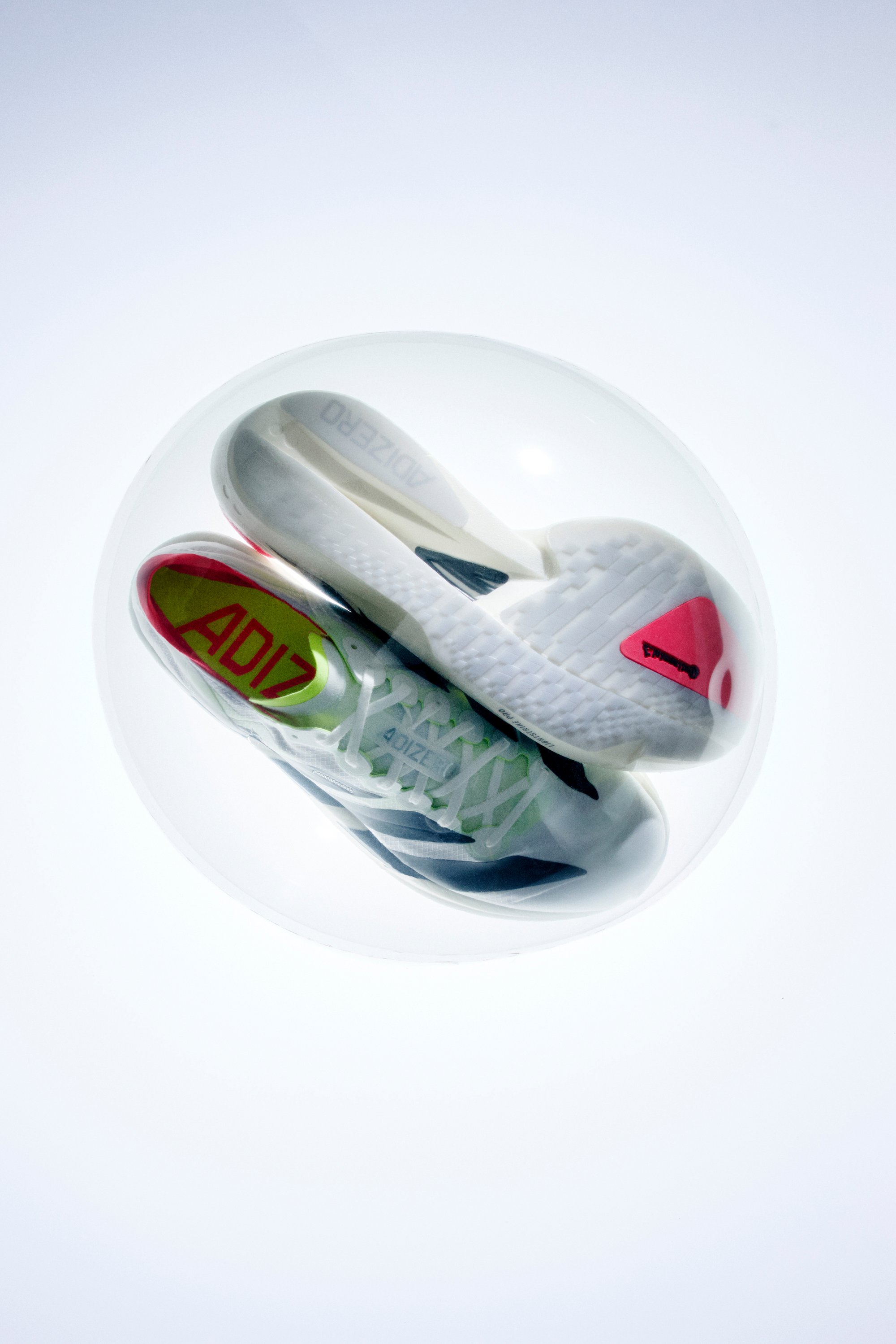 Weiße Sportschuhe mit grünen und roten Akzenten, ausgestattet mit einer strukturierten Sohle und auffälligem Branding, in einer transparenten Kugel präsentiert.