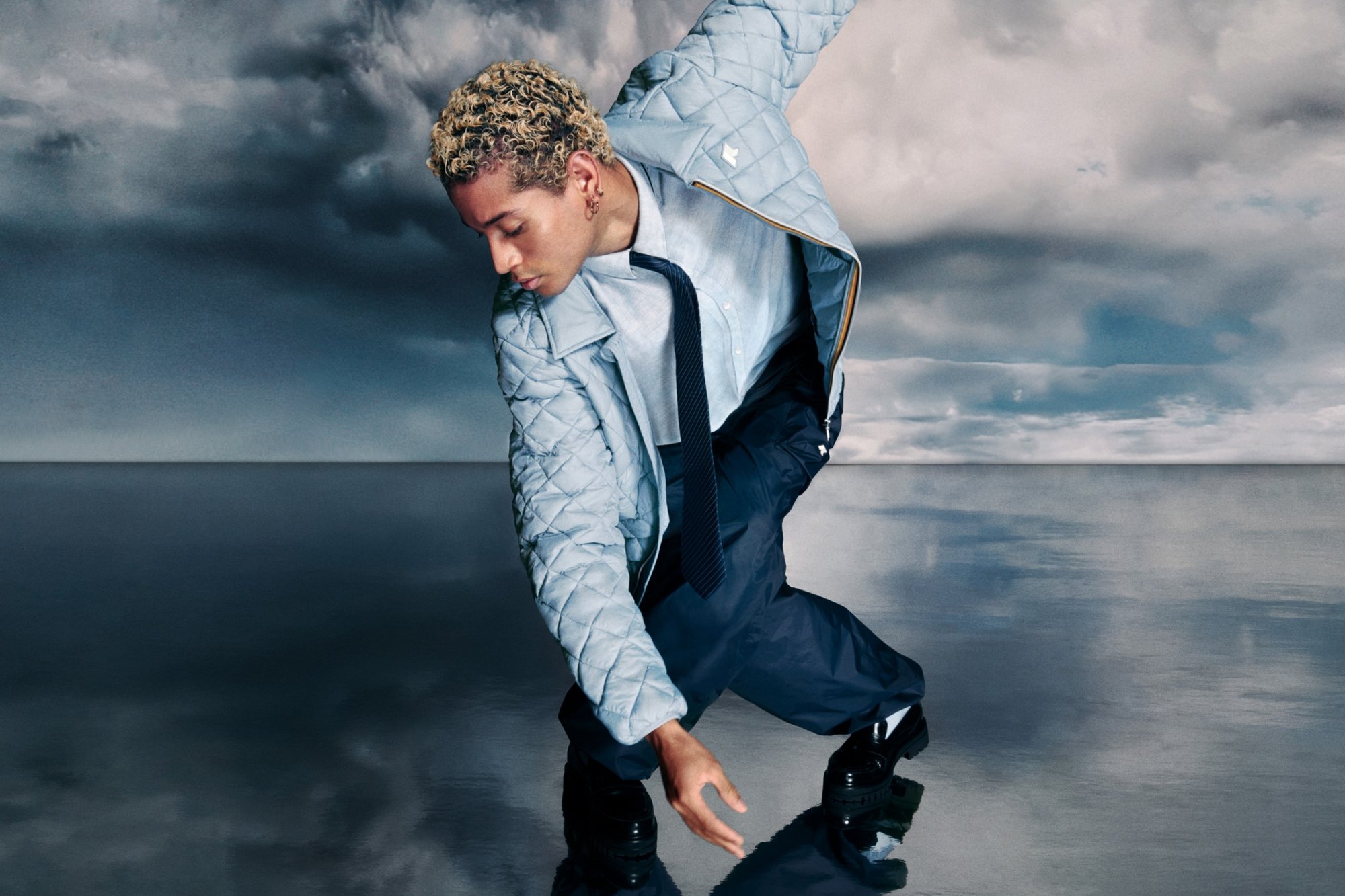 Una persona con capelli biondi ricci indossa una giacca leggera trapuntata blu chiaro e pantaloni scuri, piegandosi leggermente contro uno sfondo drammatico di nuvole.