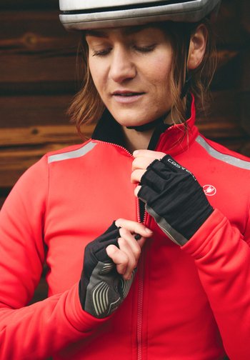 Kvinne som har på seg hvit hjelm og rød jakke, drar opp glidelåsen mens hun har på seg svarte sykkelhansker foran en trebakgrunn.