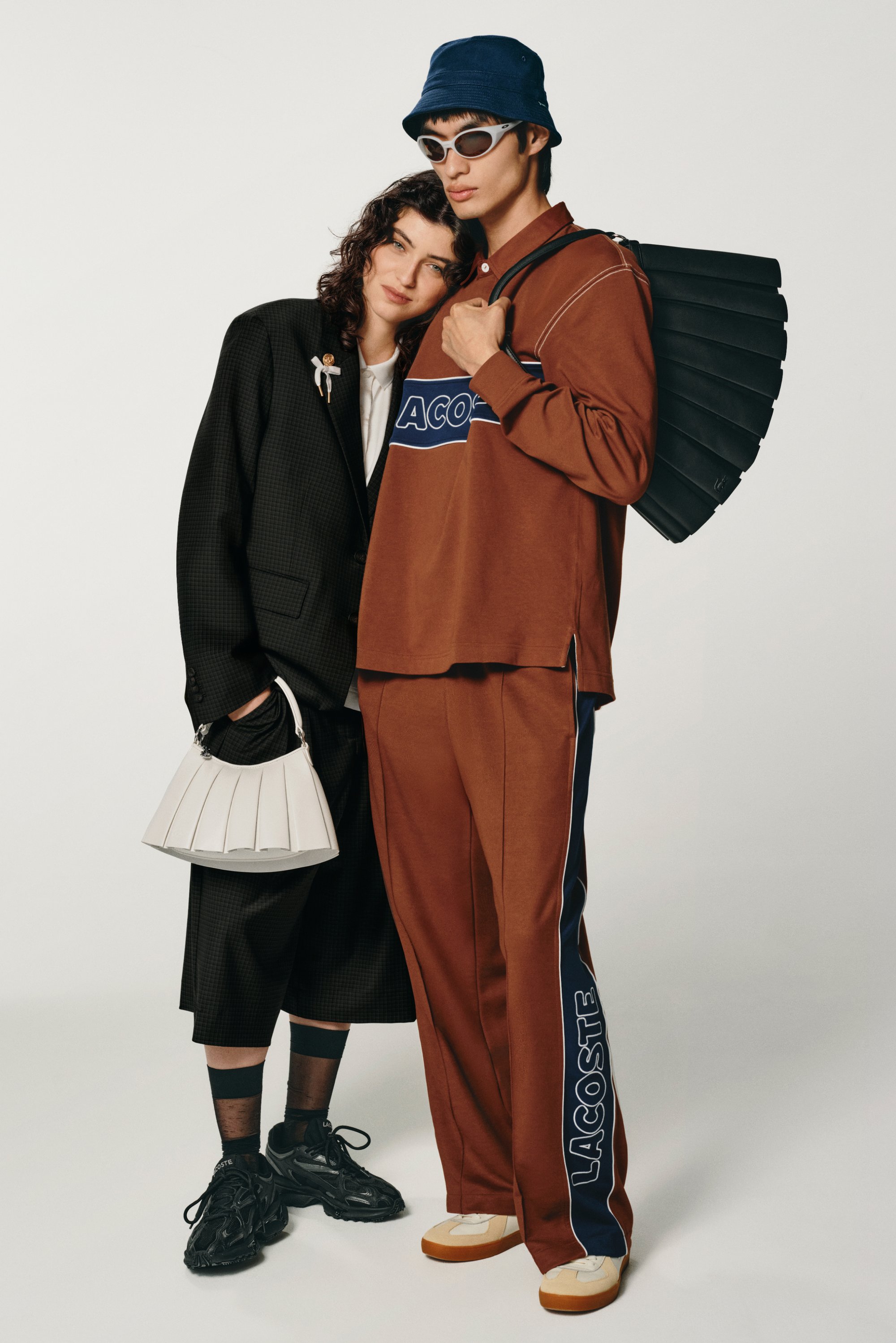 Due modelle posano vicine su uno sfondo bianco. Una indossa un completo oversize nero con una borsa bianca; l'altra è in un completo sportivo marrone e cappello a secchiello.