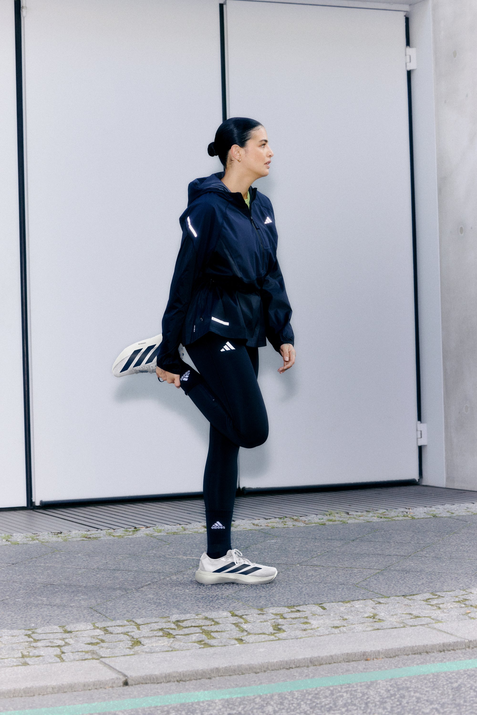 Una mujer estira su pierna mientras viste una chaqueta deportiva azul marino y leggings, preparándose para hacer ejercicio frente a un edificio moderno.