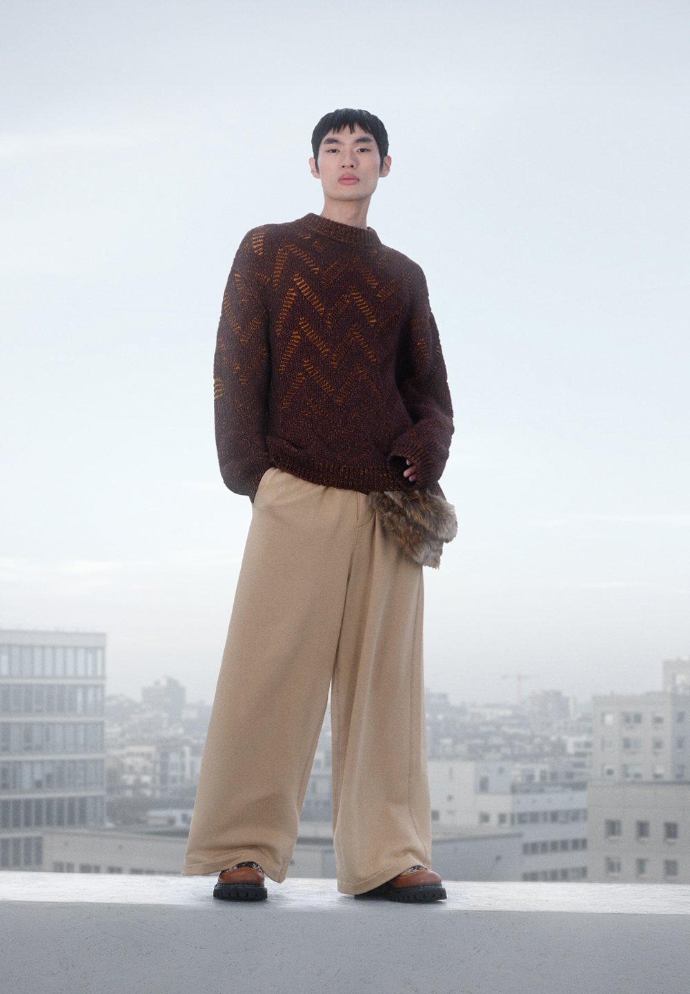 Mottled brown knit sweater with a high neck, beige wide-leg trousers, and a fur accent; paired with black and brown shoes. Urban backdrop.
