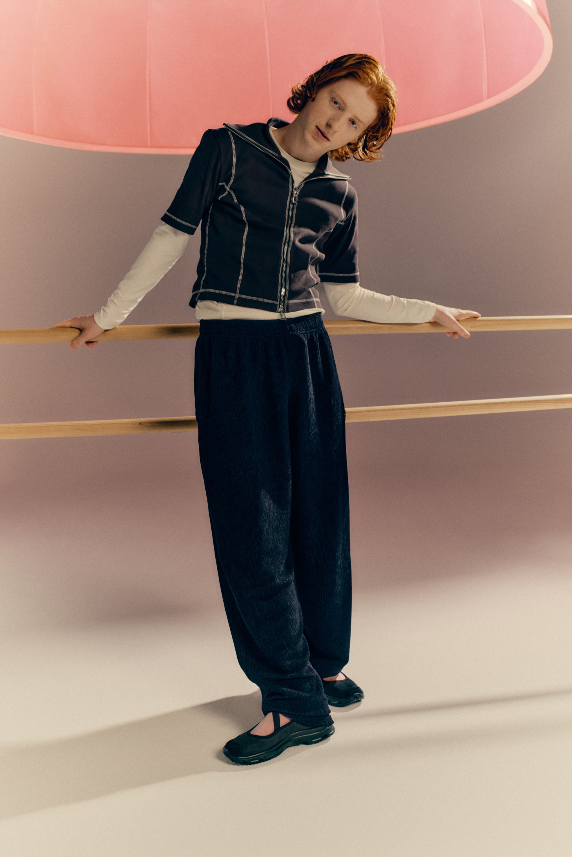 Een persoon met krullend rood haar leunt tegen een leuning, draagt een zip-up top, een lange mouw shirt en losse donkere broek, onder een roze licht.